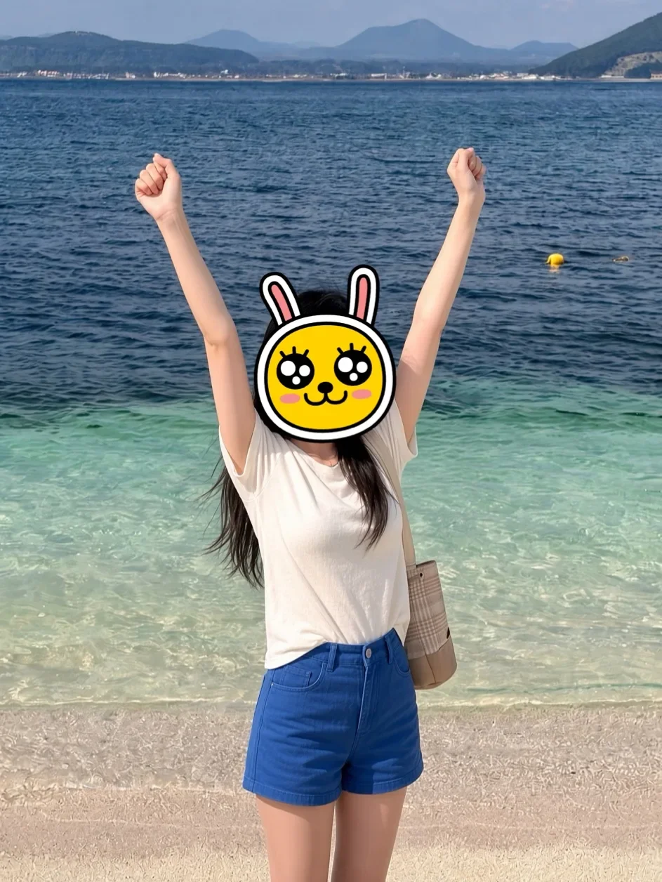 Woman standing on a white sand beach on Udo island with her arms raised, her face covered by a yellow rabbit emoji, with turquoise sea behind her
