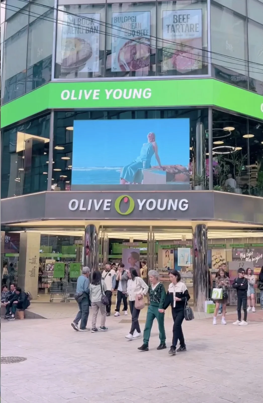 Olive Young storefront with green and white signage, a large LED screen above the entrance and people walking in front